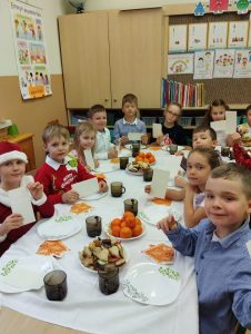 Dzieci siedzą przy stole. Na stolikach owoce, ciasteczka, dekoracje świąteczne, w dłoniach dzieci trzymają opłatki 