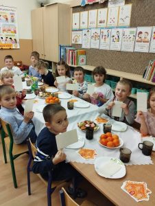 Dzieci uroczystoście ubrane siedzą przy stole, na stołach ciastka, owoce, dekoracje świąteczne. Dzieci w dłoniach trzymają opłatki.