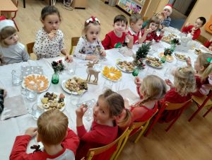 Grupa maluchy siedzi przy stole, na stołach ciastka, owoce i dekoracje świąteczne 