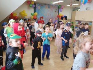 Dzieci stoją w rozsypce, przebrane za batmana. policjanta, księżniczki, tańczą kaczuszki, machają rękami.