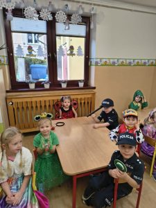 Dzieci siedzą przy stoliku w sali, w kostiumach policjantów, księżniczki, żabki, w tle okno z zimowymi dekoracjami