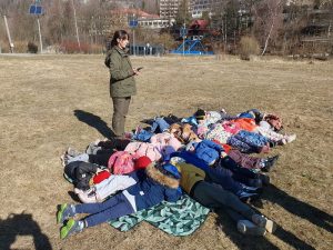 Dzieci leżą na macie, na łące, obok stoi Pani Leśnik.