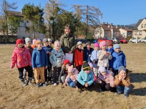 Grupa średniaki ustawiona na tle łąki, w tle drzewa i domy. Za dziećmi stoi Pani Leśnik 