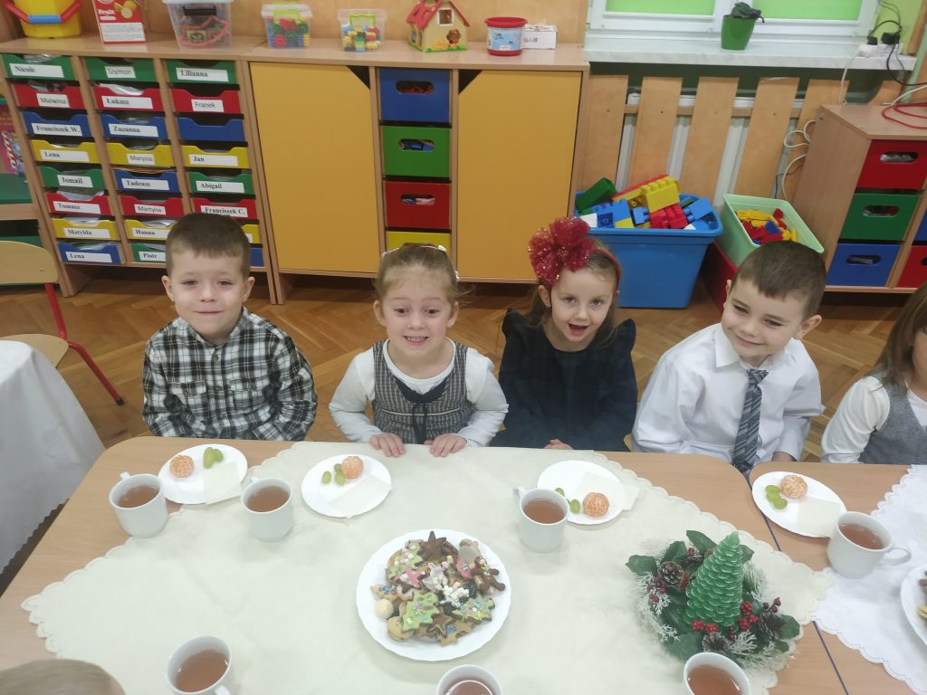Na zdjęciu znajdują się dzieci z grupy młodszej. Siedzą przy nakrytym białymi obrusami stole. Na nim znajdują się wigilijne smakołyki.