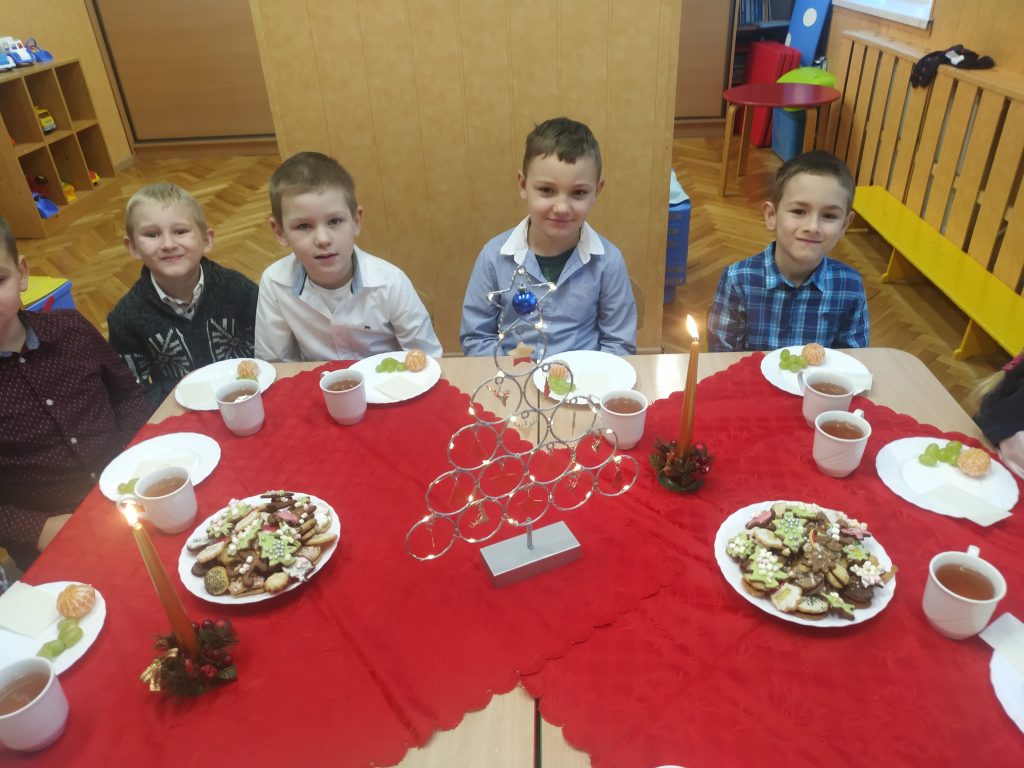Dzieci siedzą przy stole. Stół jest nakryty czerwonym obrusem. Na nim znajdują się wigilijne smakołyki.