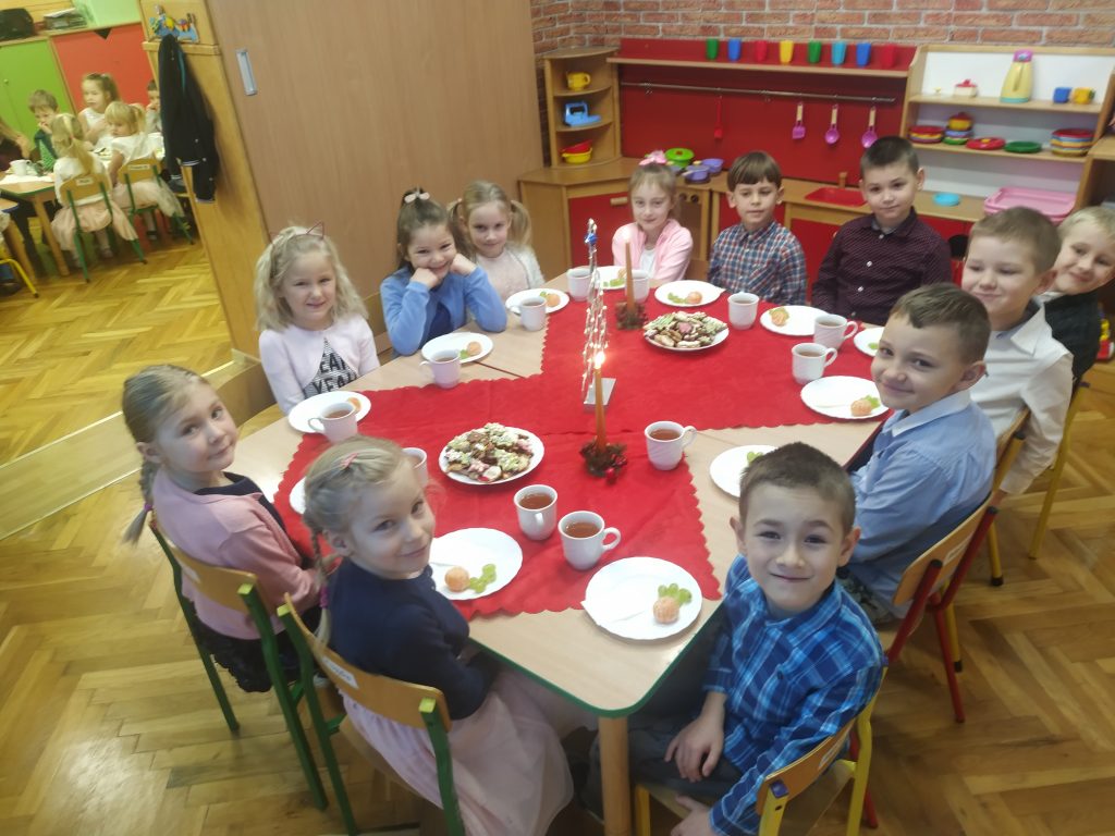 Wigilijka w grupie dzieci 5i 6-letnich. Dzieci siedzą przy nakrytym stole. Znajdują się na nim wigilijne smakołyki.