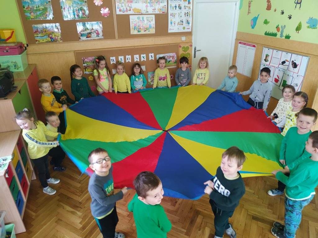 Na zdjęciu stoją dzieci w kółku i mają w ręce kolorową chustę.