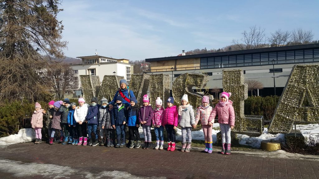 Na zdjęciu znajdują się dzieci z grupy starszej, które stoją przed szkołą wraz z swoją wychowawczynią.